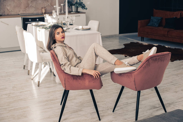 photo of a women in beige sports suit and stylish leather sneakers posing in the chair on a kitchen . selective focus - Foto, imagen