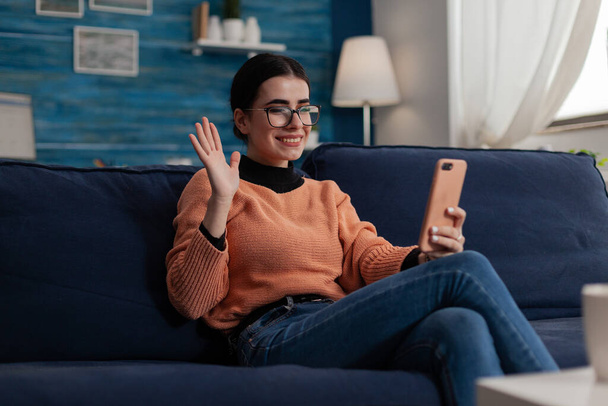 Influencer on couch holding smartphone waves hand at camera. - Foto, afbeelding