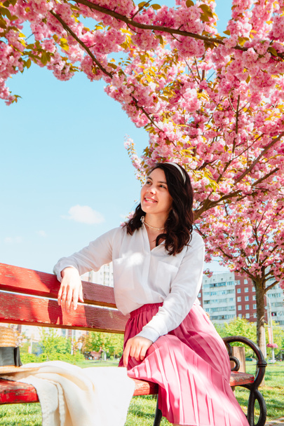 beautiful gentle woman sitting on bench under blooming sakura trees spring sunny day - Photo, Image