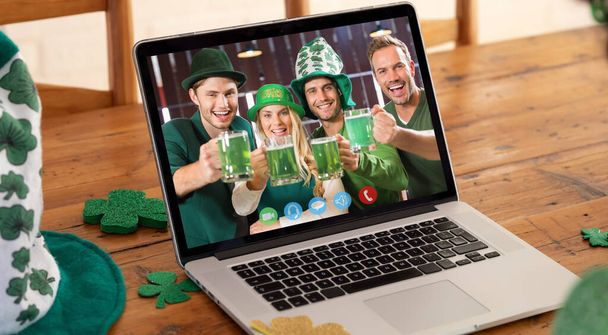 Χαμογελώντας καυκάσιους φίλους στην οθόνη του λάπτοπ κάνοντας βιντεοκλήση την ημέρα του Αγίου Πατρικίου με ποτά σε ένα μπαρ. διασκέδαση κατά τη διάρκεια του εορτασμού της γιορτής του Ιρλανδού προστάτη αγίου. - Φωτογραφία, εικόνα