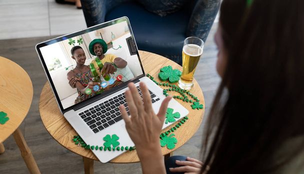 Caucasian woman at bar making st patrick's day video call waving to friends in costumes on laptop. fun during celebration of the irish patron saint's day. - 写真・画像