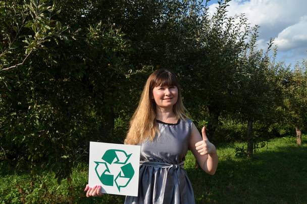 Uśmiechnięta młoda kobieta w szarej sukience trzymająca zielony znak recyklingu i pokazująca kciuk na naturalnym tle. Koncepcja ratowania planety, recyklingu i ponownego wykorzystywania śmieci. - Zdjęcie, obraz