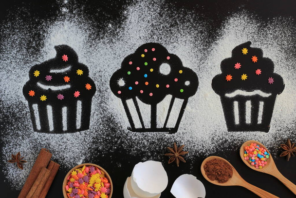 Muster von Kuchen oder Muffins aus Mehl auf schwarzem Hintergrund. Lebensmittelzutaten zum Backen - Mehl, Eier, Zucker, Kakaoanis und Süßwarenbelag. Flache Lage. - Foto, Bild