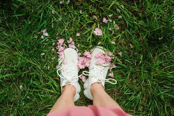 женские ноги в белых кроссовках на зеленой траве с розовыми цветущими цветами сакуры копировать пространство - Фото, изображение