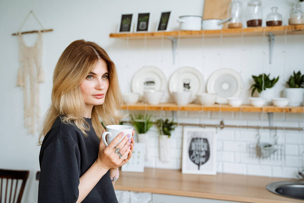 Beautiful young girl with a mug in her hands. Drink hot tea at home in the kitchen. Hold the cup in your hand to look away. Cozy interior of the dining room. High quality photo - Foto, Bild
