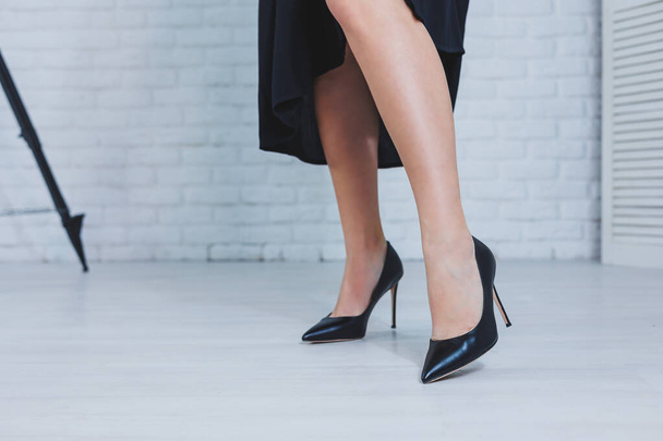 Ritratto ravvicinato di gambe femminili con tacchi alti. Una signora in abito nero si siede con lunghe gambe sottili in scarpe nere con tacco alto. Scarpe classiche da donna - Foto, immagini