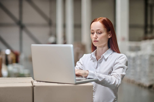 Vrouw werkt op laptop staan in de buurt van dozen in magazijn - Foto, afbeelding