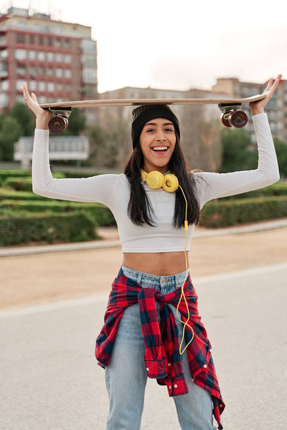 Latina woman in casual clothes posing with skateboard on her head - Foto, imagen