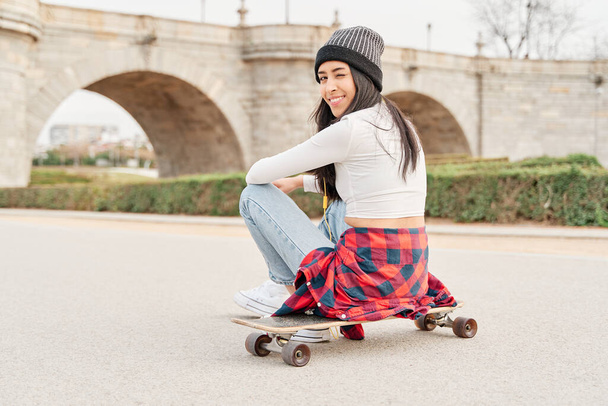 Retrato de una joven con la espalda vuelta, con gorra, camisa atada a la cintura y jeans, sentada en un monopatín en medio de un camino. - Foto, Imagen