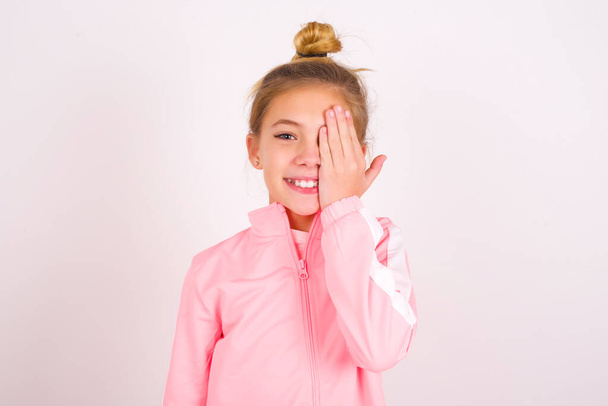 καυκάσιος κοριτσάκι φορώντας αθλητικά ρούχα πάνω από λευκό φόντο καλύπτει το ένα μάτι με το χέρι της, αυτοπεποίθηση χαμόγελο στο πρόσωπο και έκπληξη συγκίνηση. - Φωτογραφία, εικόνα