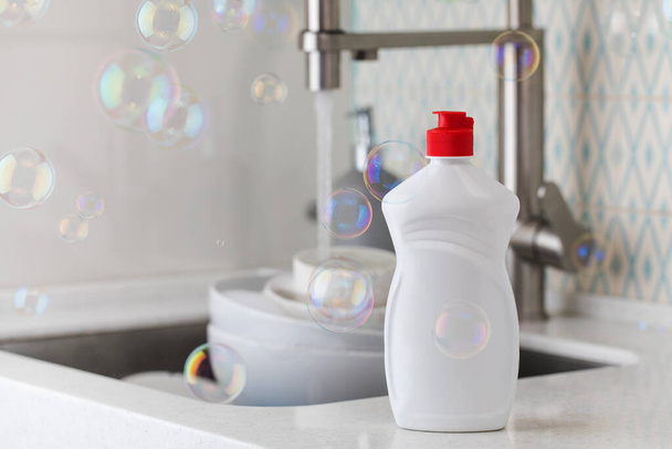 White bottle with dishwashing gel with soap bubbles around on the background of a sink with dirty dishes. - Fotografie, Obrázek