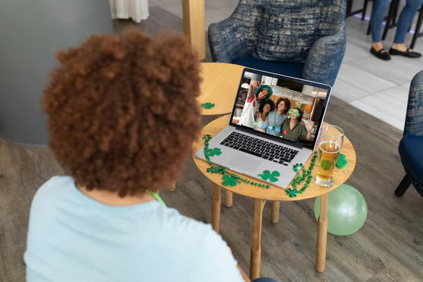 Mixed race man at bar making st patrick's day video call to friends on laptop. fun during celebration of the irish patron saint's day. - Foto, Imagen