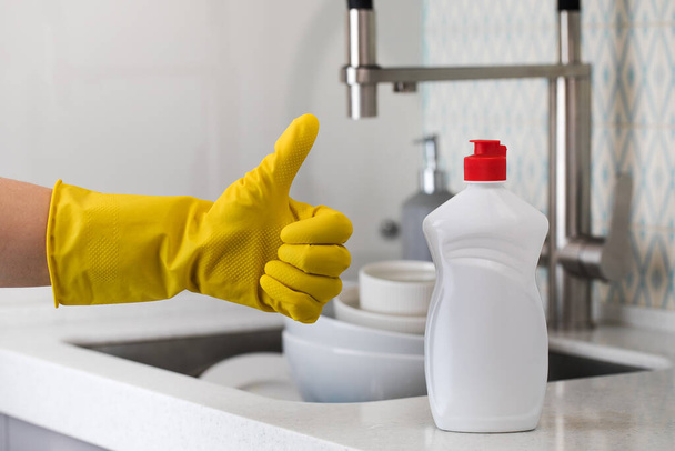 A white bottle with dishwashing gel and a hand in a yellow rubber glove waggles its thumb up on a sink with ddishes. - Foto, afbeelding