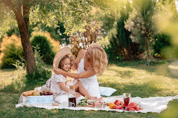 A beautiful blonde girl with curly hair and her daughter had a picnic in nature on a hot sunny day. Family on a picnic, vegetables, fruits, juice and baguette. Selective focus, defocus. - Foto, Bild