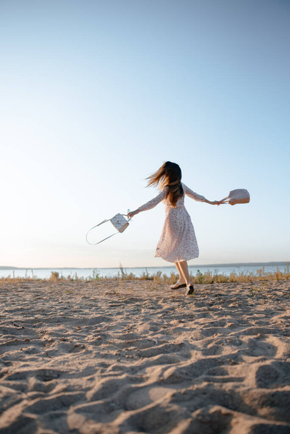 Kaunis tyttö pitkät paksut hiukset valkoisessa mekossa kävelee pitkin merenrantaa aurinkoisena kesäpäivänä, kaksi valkoista pussia käsissään. Onnen ja rentoutumisen käsite. Kävele pitkin merta. - Valokuva, kuva