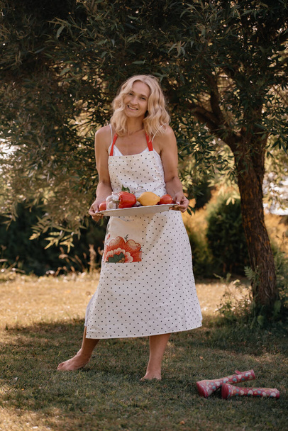 Beautiful happy woman with blond curly hair in a long white apron holding a tray with fruits and vegetables in her hands while standing outdoors. Summer picnic. - Foto, afbeelding