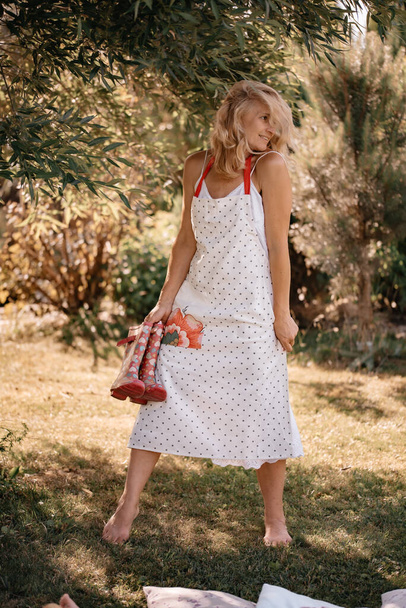A beautiful woman with blond curly hair in a long white apron is resting in nature on a sunny summer day, a girl with rubber boots in her hands. Happiness and relaxation concept. - Foto, Bild