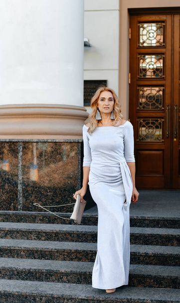 A beautiful blonde girl in a long tight white dress with a clutch in her hands is standing on the stairs near an architectural building, in the background there is a large wooden door. Styles and fashion concept. - Foto, Bild