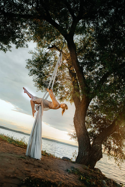 A young attractive girl in a white dress goes in for sports on nature near a tree, against a background of clear sky. Health and sport concept. Aerial gymnastics. - Foto, afbeelding