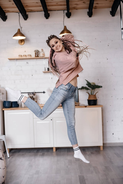 A young smiling girl in blue jeans and a pink sweater with a hood is dancing in the kitchen. Girl with afro braids. Photo in motion. - Foto, immagini