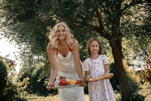 木の背景に果物や野菜を手に白いドレスを着た少女と彼女の母親。自然の中で夏のピクニック。母と娘は公園で一緒に楽しんでいます. - 写真・画像