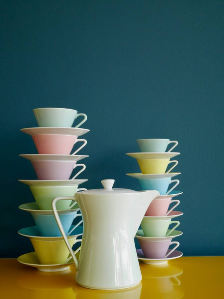 Stack of colorful vintage tea and coffee cups and tea pot on yellow table against petrol background. Vertical. High quality photo - Photo, Image