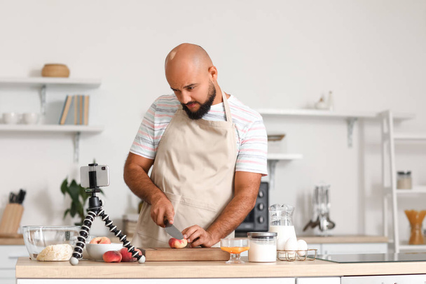 Schöner Mann schneidet Pfirsich, während er Kochvideo-Anleitung in der Küche verfolgt - Foto, Bild