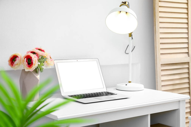 Moderno lugar de trabajo con portátil, flores en jarrón y lámpara brillante cerca de la pared de luz - Foto, Imagen