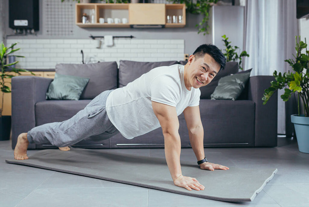 Portrait of an Asian man at home, doing fitness and sports, at home in the living room, on a sports mat, pushing off the floor smiling and looking at the camera - Photo, Image