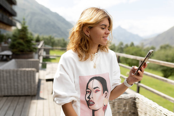 Close up outside portrait of blond charming woman with wavy hair wearing white pullover is holding smartphone and smiling in sunlight with mountains view. High quality photo - Photo, Image