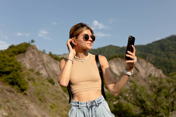 Atrakcyjny cute girl z zebranych włosów w okularach przeciwsłonecznych w beżowym t-shirt robi selfie przeciwko górom w świetle słonecznym. Szczęśliwa kobieta podróżująca w górach w ciepły wiosenny dzień. Wysokiej jakości zdjęcie - Zdjęcie, obraz