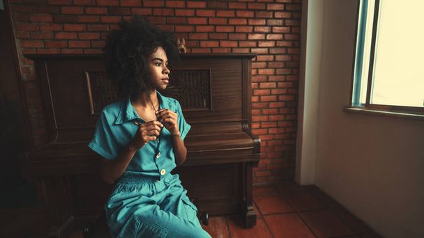 View of a ravishing young Brazilian female with curly afro hair sitting on the chair in the room and pensively looking outside the window with a piano behind her and a copy space place on the right - Foto, immagini