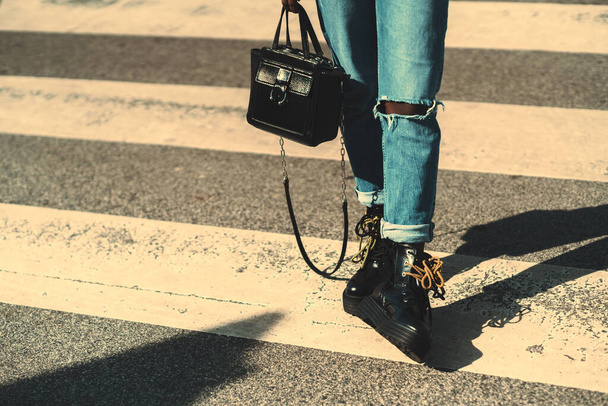 Close-up view of female legs in ripped jeans and with black glossy leather boots with yellow laces, standing on the zebra pedestrian crosswalk on the road with an elegant bag in the hand, a sunny day - Fotoğraf, Görsel