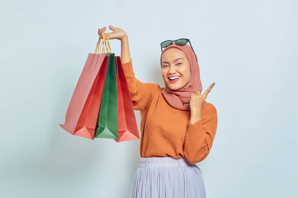 Весела красива азіатська мусульманка в коричневому светрі і окулярах тримає сумку для покупок, вказуючи на копіювання простору пальцем ізольованим на білому тлі. Всесвітній день прав споживачів
 - Фото, зображення