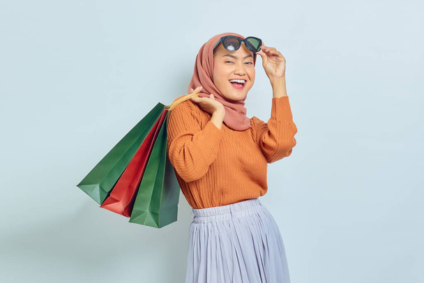 Portret van opgewonden jonge Aziatische moslim vrouw in bruine trui zonnebril houden pakket tas met aankopen na het winkelen geïsoleerd op witte achtergrond. Werelddag van de rechten van de consument - Foto, afbeelding