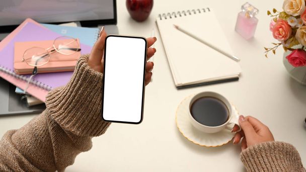Entspannte Frau im gemütlichen braunen Pullover genießt ihren morgendlichen Kaffee und schaut sich am Schreibtisch ein paar Online-Inhalte auf dem Smartphone an. Telefon weiße Bildschirm-Attrappe. - Foto, Bild