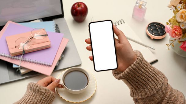 Freelancerin oder Bloggerin im gemütlichen braunen Pullover liest auf ihrem Smartphone einen Online-Inhalt oder Weblog und nippt am morgendlichen Kaffee an ihrem Tisch. - Foto, Bild