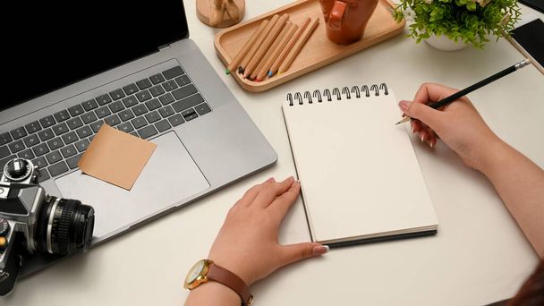 Kobieta blogerka podróży pisze recenzje podróży na spiralnym notatniku w nowoczesnym biurze. Laptop, kamera i akcesoria na stole. - Zdjęcie, obraz