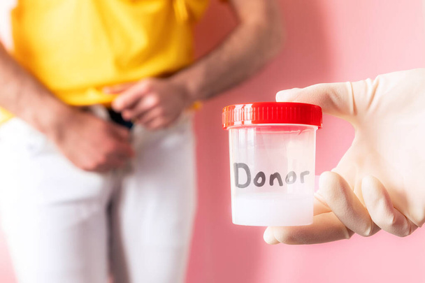 Doctor's hand in a medical glove holds a plastic jar with a sample of sperm. In the defocused background a man shyly zips up his jeans. The concept of sperm donation and artificial insemination. - Фото, зображення