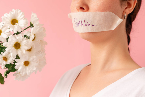 Close up of caucasian woman's face on a pink background with a taped mouth and a bouquet of chamomiles. The concept of March 8 and women's secrets. - Foto, imagen