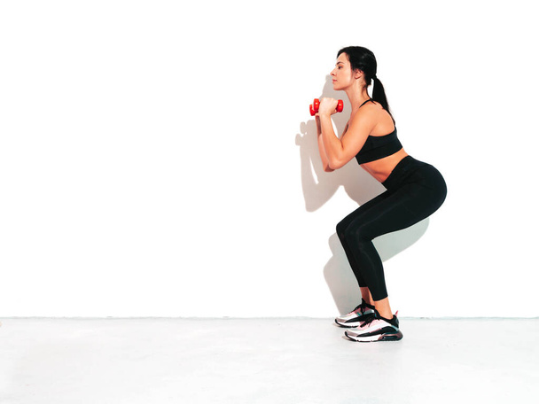 Fitness confident woman in black sports clothing. Sexy young beautiful model with perfect body. Female isolated on white wall in studio. Stretching out before training.Making squats with dumbbells - Foto, afbeelding