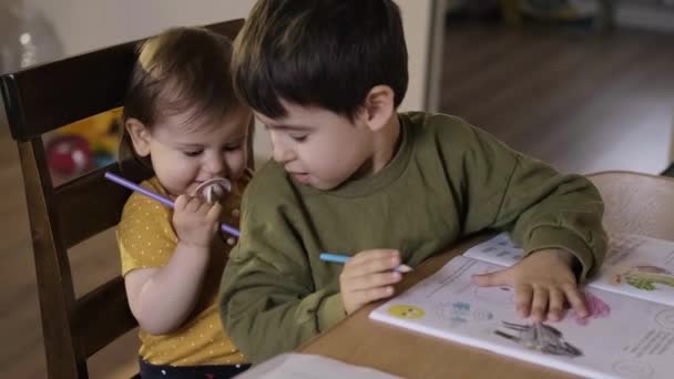 Hermano jugando con la hermanita mientras disfrutan del tiempo libre juntos en casa dibujando con lápices de colores mientras están sentados en la mesa. Educación infantil. Cara feliz - Metraje, vídeo