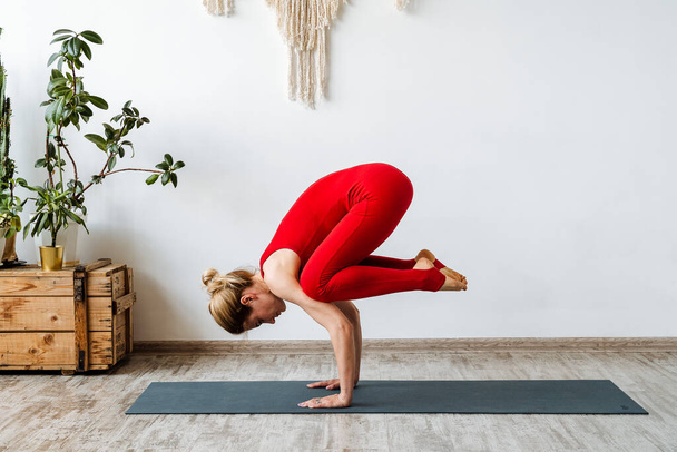 A young beautiful white girl makes kakasana on a yoga mat. Home sports stretching strength exercise standing on your hands. High quality photo - Foto, Imagen