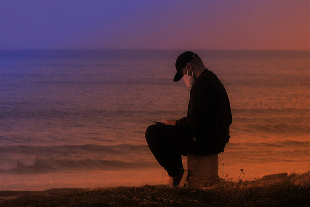 Uomo in nero seduto su una roccia che legge al tramonto - Foto, immagini