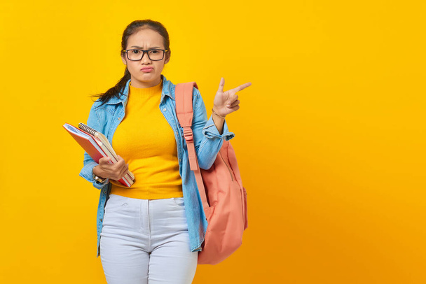 Portrait of confused young Asian woman college student in denim clothes with backpack holding notebook and pointing finger at copy space isolated on yellow background - Φωτογραφία, εικόνα