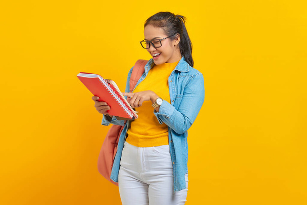 Lächelnde Studentin in Jeanskleidung mit Rucksack, die im Arbeitsheft auf gelbem Hintergrund liest. Ausbildung im Hochschulkonzept der High School - Foto, Bild