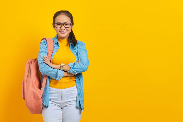Jeune étudiant asiatique souriant en tenue denim avec sac à dos traversé la poitrine et l'air confiant isolé sur fond jaune. Éducation dans le concept de collège universitaire du secondaire - Photo, image