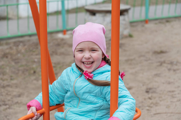Un niño con una chaqueta azul oscila en un columpio. Feliz niña de cinco años con coletas divirtiéndose en el patio de recreo - Foto, imagen