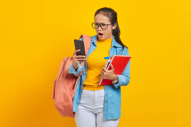 Portrait of surprised young Asian woman student in denim clothes with backpack using mobile phone typing sms message and hold books isolated on yellow background - Photo, Image