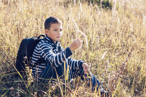 Autumn portrait of teenager boy sitting on grass. Young man with backpack resting relaxing, contemplating, thinking, daydreaming. Teen deep in thought Local travel. Active lifestyle People from behind - Zdjęcie, obraz